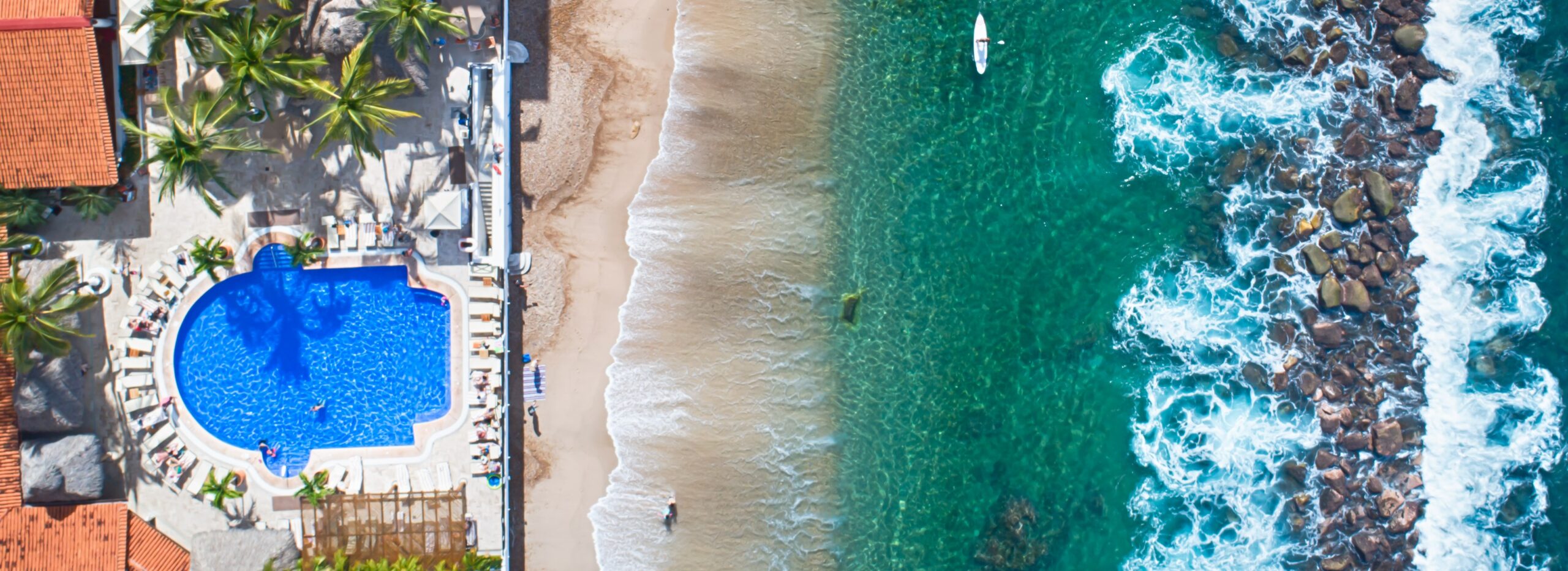 Aerial view of a beachfront resort with a large pool, palm trees, and a sandy beach. The ocean is a vibrant turquoise color with waves crashing on rocky shores. A person is paddleboarding in the distance.